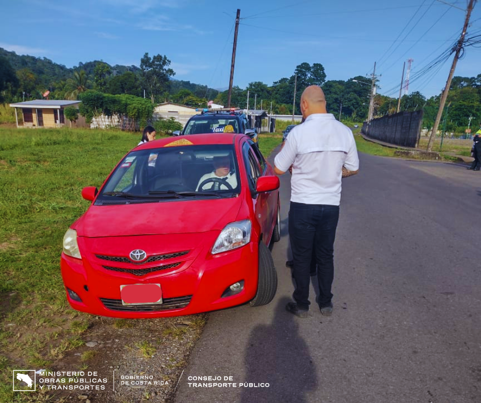 Taxi rojo estacionado a un costado de la carretera y compañeros del CTP hablando con chofer.